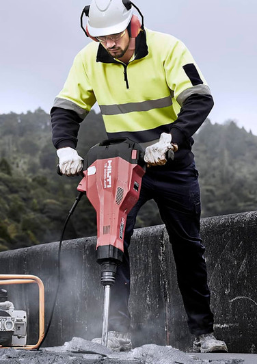 A worker in a fluorescent yellow and black hooped pullover operates a demolition tool at a construction site.