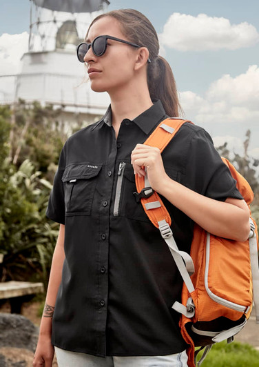 A woman in a black short sleeve outdoor shirt carries an orange backpack, standing near a lighthouse.