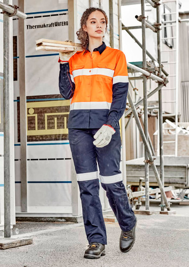 A woman wearing orange and navy workwear, featuring a utility shirt and pants, stands on a construction site holding timber.