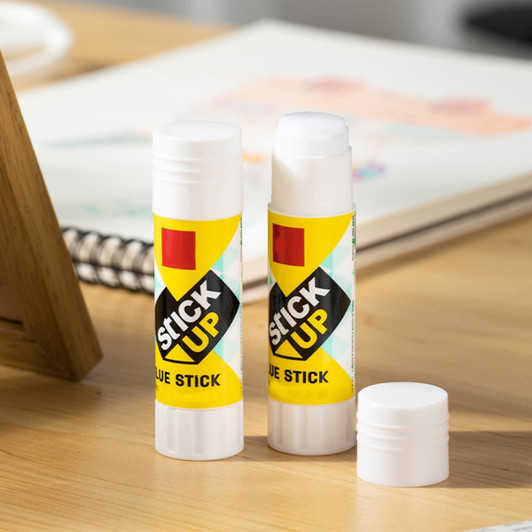 A glue stick in a bright yellow and green design, with one cap removed, placed on a wooden desk.