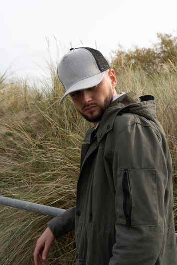A grey marle cap with a mesh back worn by a man in a green jacket, standing near grassy terrain.