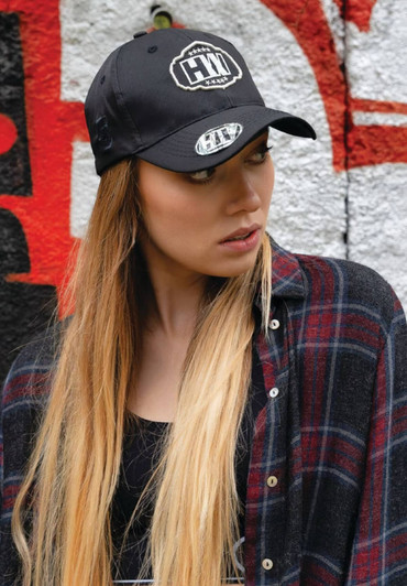 A black cap with a logo is worn by a model with long hair, against a graffiti backdrop.
