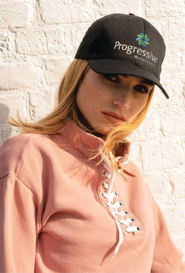 A black organic brushed heavy cotton cap with a logo, worn by a woman in a pink sweatshirt against a white wall.