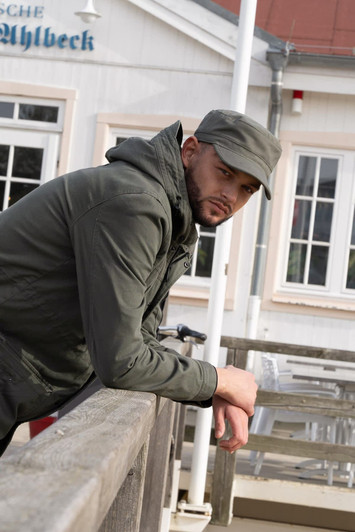 A man wearing an olive green Sports Twill Military Cap leans against a railing, dressed in a jacket. The background features a building.