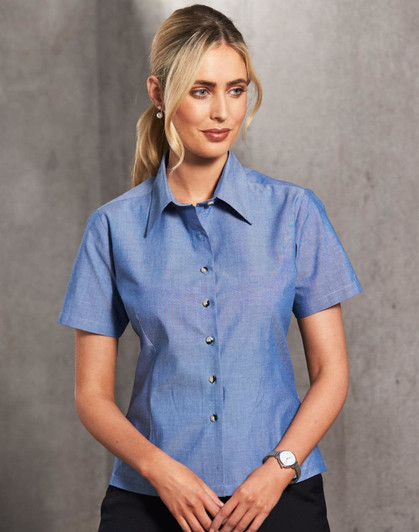 A button-up ladies' short sleeve shirt in chambray blue, featuring a collar and a row of buttons, with a logo.