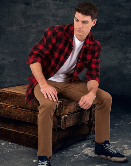 A man sits on a wooden crate wearing a red and black plaid flannel shirt over a white t-shirt with beige pants.