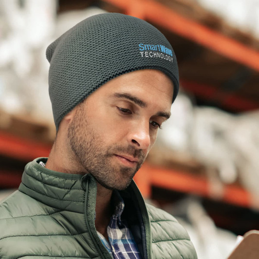 A grey garter knit beanie worn by a man, featuring a logo, set against a blurred warehouse background.