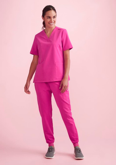 A model wearing women's pink jogger scrub pants and a matching pink scrub top, set against a light pink background.