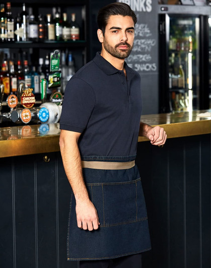 A man wearing a denim half apron stands at a bar, featuring a logo on the apron and a dark background with shelves of drinks.