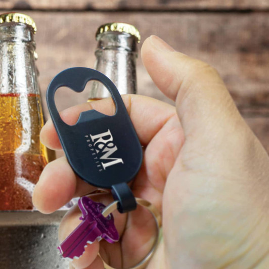 A drink bottle opener key ring in black, held in a hand, with glass bottles in the background. It features a logo.