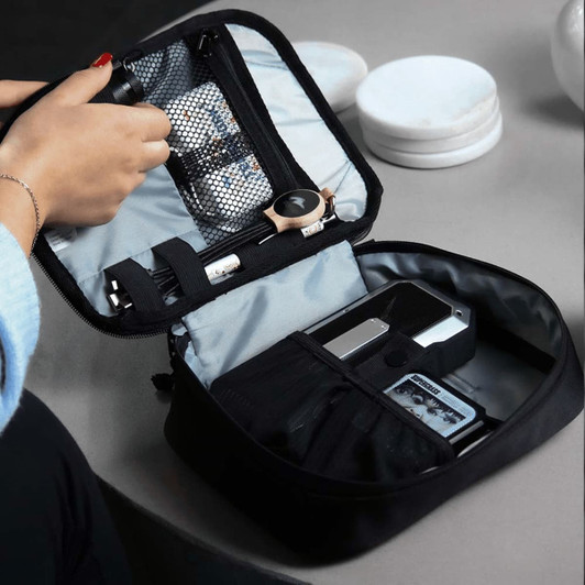 A black tech case opened to reveal various compartments holding gadgets and accessories. The case features a mesh pocket and organisers.