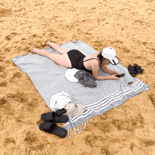 A grey and white striped throw blanket lays on the sand, with a person lounging on it, surrounded by clothing and accessories.