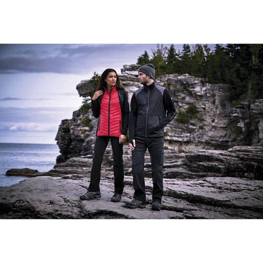 A woman in a red insulated jacket and a man in a black jacket stand on rocky terrain near a body of water, surrounded by trees.