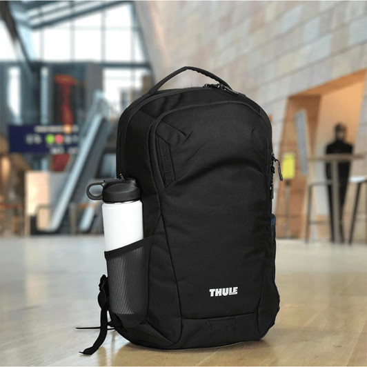 A black Thule Recycled Lumion 16" computer backpack with a water bottle in the side pocket, set in a modern interior space.