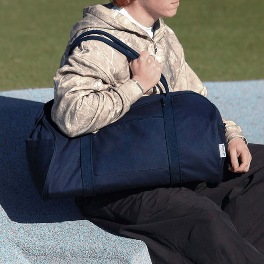A person seated on a surface holds a large navy duffel bag with two sturdy handles. The bag has a simple design and branding.