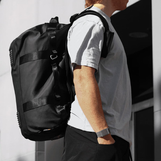 A black duffel bag is worn on one shoulder by a person in a casual outfit, set against a building backdrop.