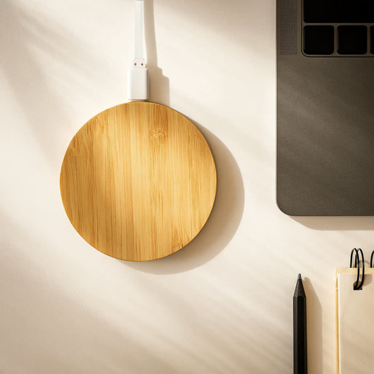 A round bamboo wireless charger in a natural finish, placed beside a laptop and stationery on a desk.
