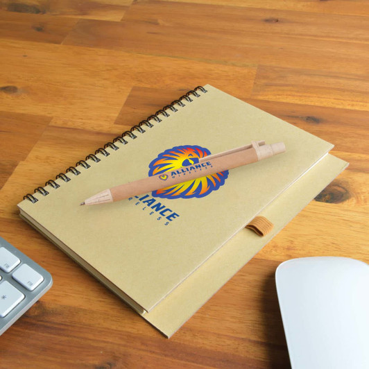 A beige spiral notebook with a logo and a wooden pen resting on a wooden surface alongside a keyboard and mouse.