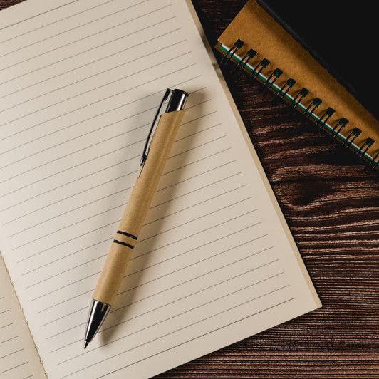 A pen with a brown paper body and silver accents rests on a lined notepad with a spiral notebook nearby.