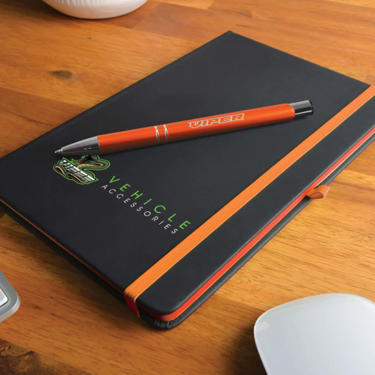 A black notebook with an orange strap and an orange pen resting on top, featuring a logo.