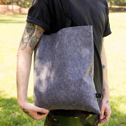 A grey RPET felt tote bag held by a person, set against a green outdoor background. The bag features a logo.