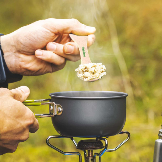 A hand holds a light-colored wooden utensil over a steaming gray pot, set against a natural background.