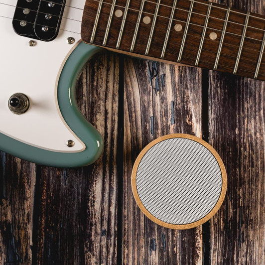 A round white Bluetooth speaker with a wooden base, placed beside a teal electric guitar on a rustic wooden surface.