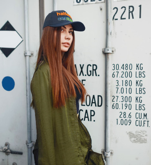 A woman with long hair wears a navy cap with a logo, standing next to a shipping container. She is dressed in an olive green jacket.
