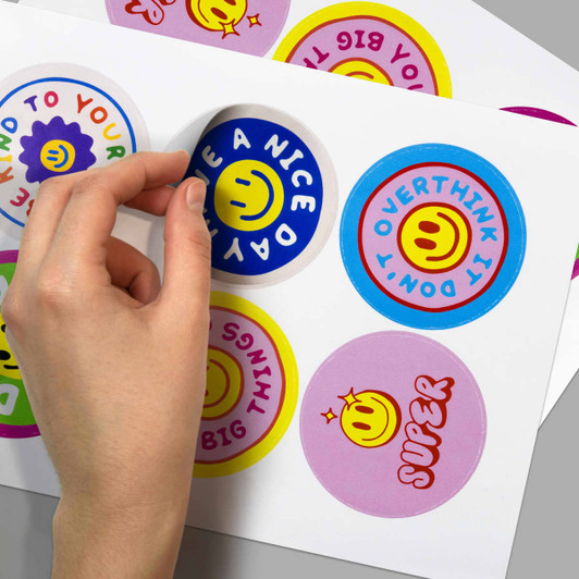 A hand peels a glossy sticker from a sheet featuring colourful circular designs with smiley faces and text.