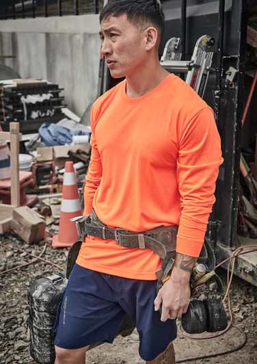 A man wearing an orange long sleeve high visibility tee is standing at a construction site, equipped with a tool belt.