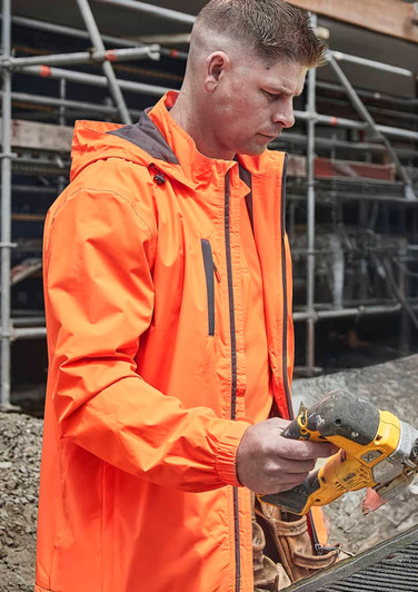 A man wearing an orange unisex packable rain jacket, focused on using a power tool on a construction site.