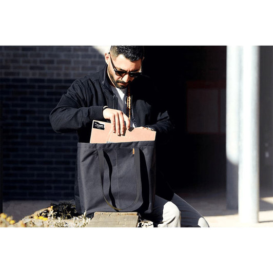 A black tote bag with long handles is being carried by a person. The bag contains a notebook and has a logo.