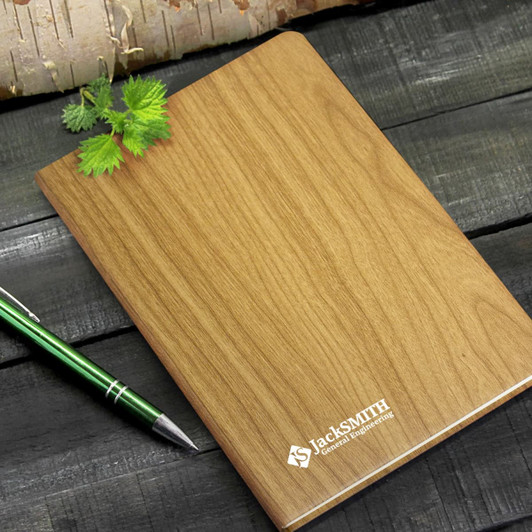 A wooden finish notebook with a logo, placed beside a green pen and a leaf on a rustic wooden surface.