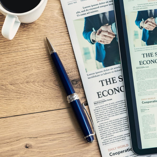 A blue ballpoint pen with a silver trim resting on a wooden table alongside a newspaper and a coffee cup.