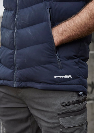 A navy unisex hooded puffer vest with a zip pocket and a logo on the side, worn over grey cargo pants.