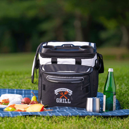 A black and white rolling cooler with a logo, placed on grass near picnic food and drinks.