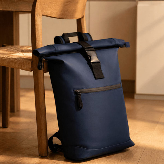 A navy roll-top backpack with a front zip pocket rests against a chair in a warm, indoor setting.