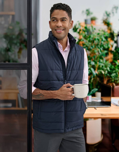 A man wearing a navy insulated puffer vest stands holding a mug, with greenery in the background.