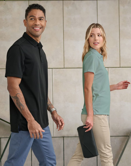 A black men's polo and a green women's polo are worn by two people against a neutral background. Both garments have logos.