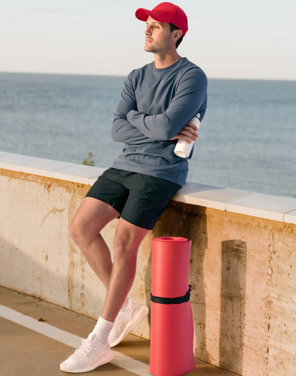 A man in a grey long-sleeve tee and red cap stands by a beach, holding a drink bottle next to a red yoga mat.