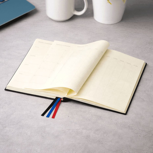 A black diary with cream pages opened to reveal weekly layouts, featuring coloured ribbon bookmarks.