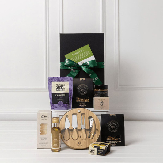 A gourmet gift pack featuring a wooden serving board, assorted gourmet snacks, olive oil, and a jar of jam, all in a black box.