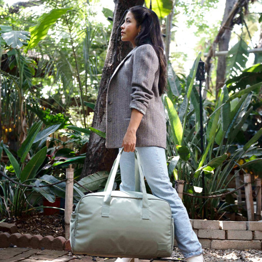A duffle bag in a light green shade is held by a person in a garden setting, surrounded by lush foliage.