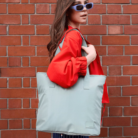 A woman with long hair holds a light blue tote bag against a brick wall, wearing a red jacket and sunglasses.