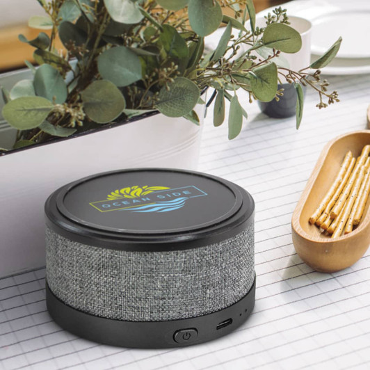 A wireless speaker charger with a grey fabric exterior and a black base, placed beside greenery and snacks.