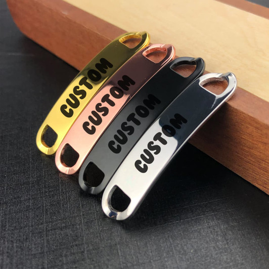 Four Smart Shoelace Charms in gold, rose gold, black, and silver, featuring a logo, arranged on a wooden surface.