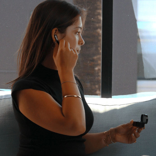 A woman wearing black Prixton TWS157 earbuds, holding the charging case, seated by a window.