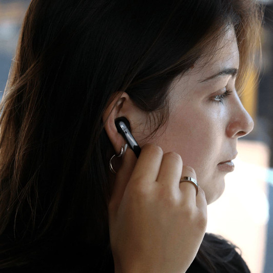A woman is wearing black Prixton TWS157 earbuds in her ear, adjusting one while looking off to the side.