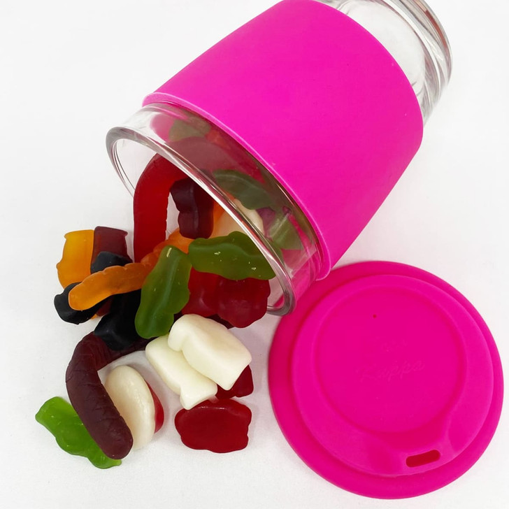 A reusable glass coffee cup with a pink silicone sleeve, filled with assorted jelly lollies spilling out.