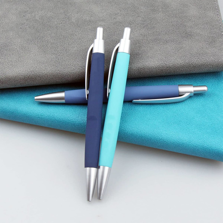 Three plastic pens in navy blue and light blue resting on textured grey and turquoise surfaces, featuring metallic accents.
